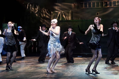 Viola in silvery flapper dress in the middle of a line of women dancing, with men in suits dancing behind. The women beside Viola are in black dresses, with the one to the left sporting a white scroll pattern on the front and a white headpiece.